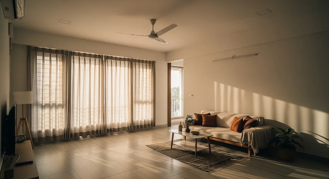 Sunlit, tidy living room in a Chennai apartment with clear floors, cotton curtains, and a ceiling fan